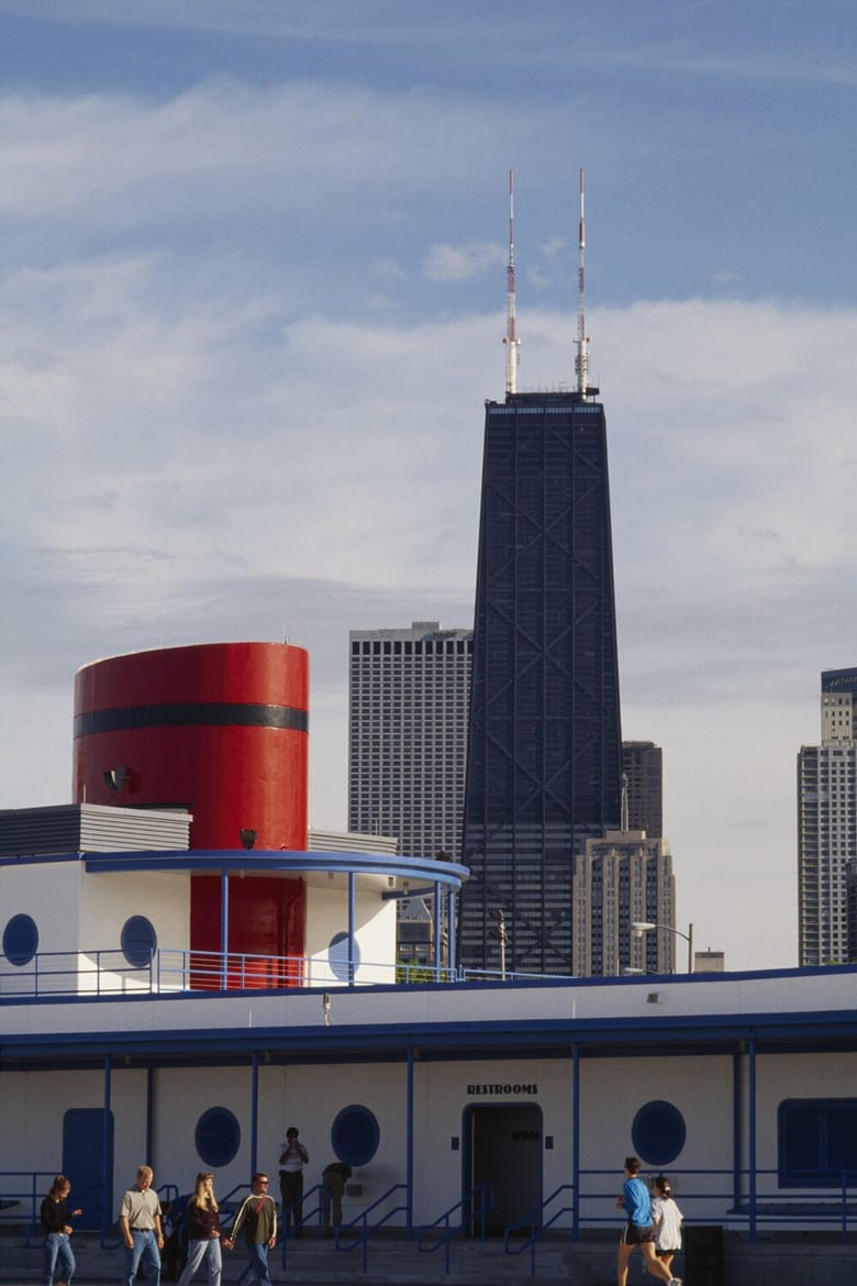 A ground-level shot frames the John Hancock Center (now 875 North Michigan Avenue) in the background, towering over a foreground building with a nautical design. The foreground building is white with blue trim and features circular windows resembling portholes, and a large red cylindrical structure on its roof. The word "RESTROOMS" is visible above an entrance. Several people are walking around the foreground building's patio area. The sky is partly cloudy.
