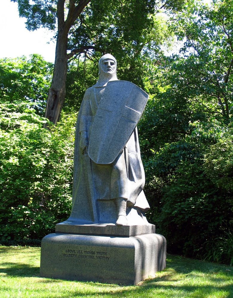 Stone statue of an armored man with a large shield with grass surrounding.