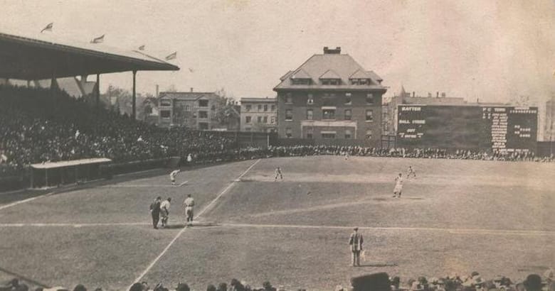 Black and white image of a baseball field and players standing on it with stands full of people on the left and in the background
