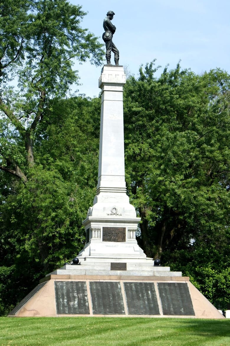 Large white monument with 12 smaller grave markers on grassy plain.