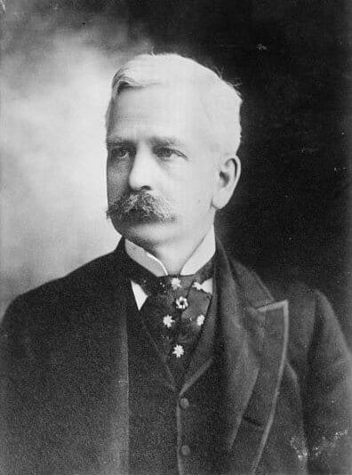 Black and white image of a man with white hair and a mustache wearing a suit and looking to his right