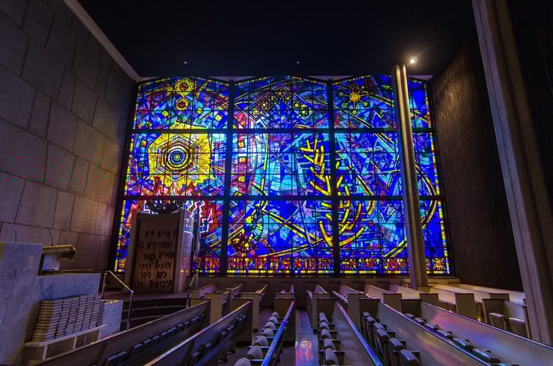Interior image of a synagogue showing pews from the side and a colorful stain glass window directly beside them 