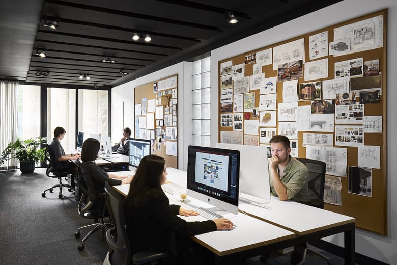 Office workers sitting at wide desks with large monitors with a corkboard wall in the background