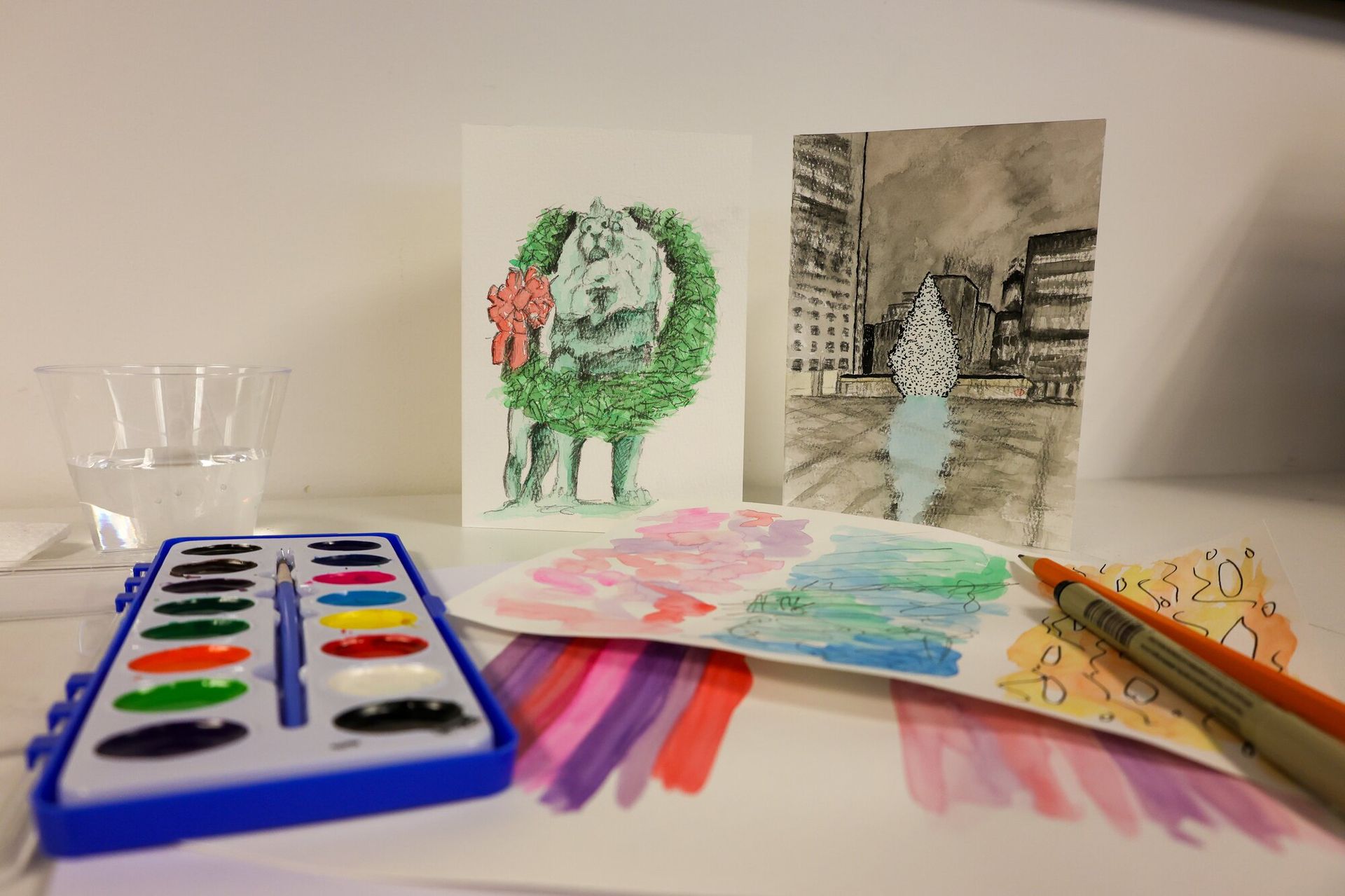 Image of some water color paintings propped up next to a paint pallet and some pens