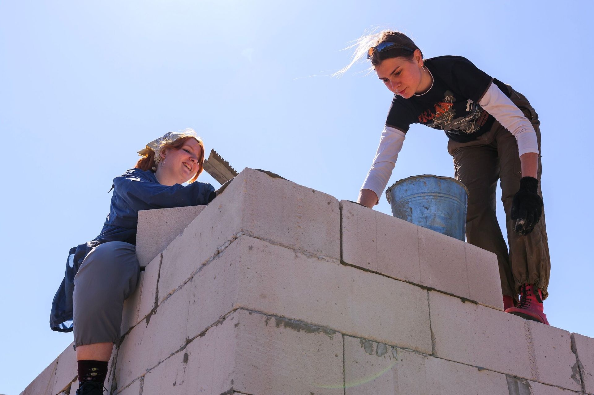 Image or two women one sitting and one standing, on top of a concrete block structure, building it. 