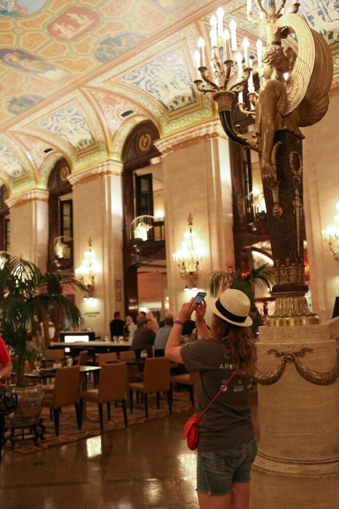 Picture of a woman taking a photo inside a hotel lobby which contains people sitting on chairs, golden light fixtures, and a painted ceiling