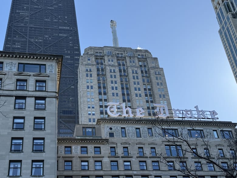 Close up image of the top of a tall building with a sign on top in white letters that reads "The Drake"