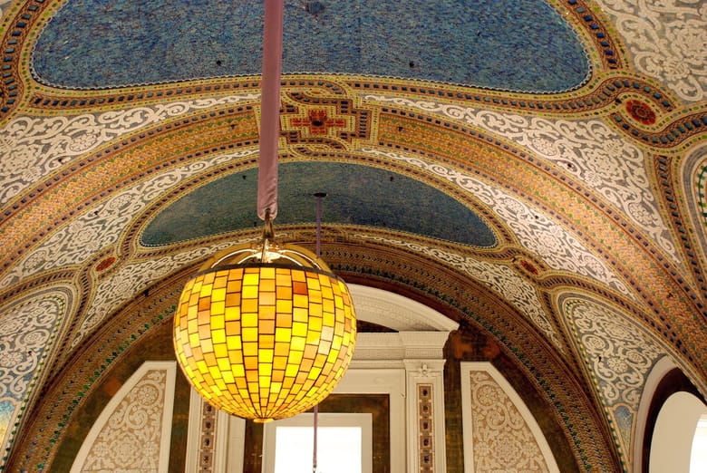 Image of a mosaic vaulted ceiling that is blue, brown, and tan with a globe shaped yellow stain glass chandelier. 