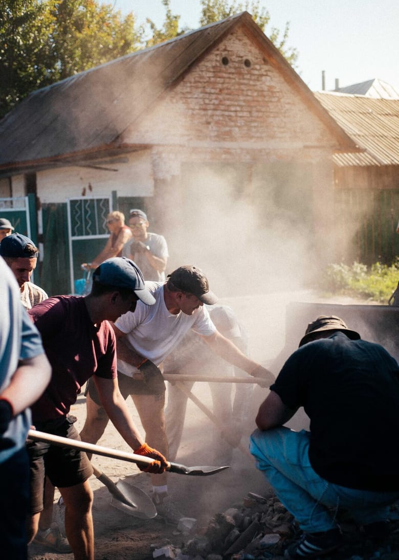 Image of several men standing, with two holding shovels. There is a white house in the background.