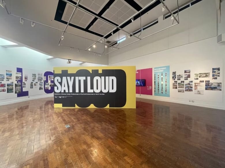 Gallery space with a wall that says "Say it loud" in large white all caps text on a black and yellow background.