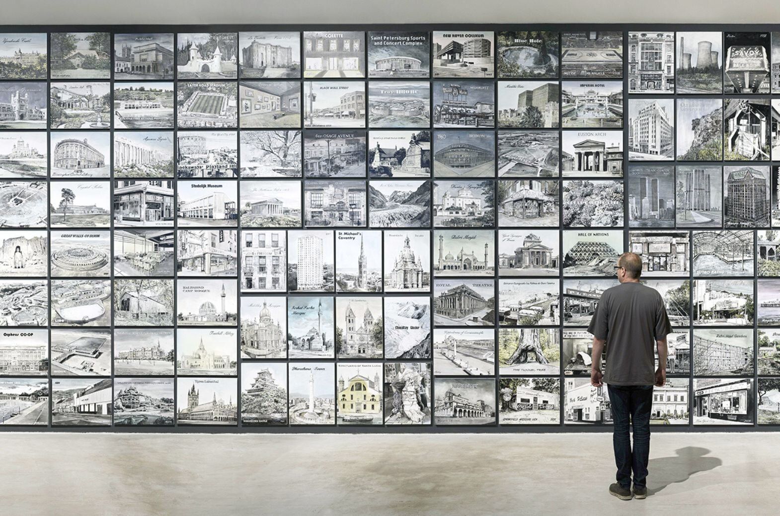 Man standing in front of a wall of paintings all the same size hung in a grid.