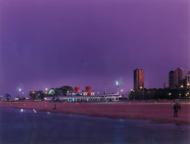  A wide-angle, twilight shot of North Avenue Beach in Chicago, bathed in a purple hue. The North Avenue Beach boathouse, with its distinctive red-topped "smokestacks" and white structure, is centrally located on the left, illuminated by scattered lights. The beach in the foreground appears damp, reflecting some of the sky's light. In the background, the Chicago skyline rises, with several tall buildings glowing with orange and white lights. Streetlights line the beach path, and trees are visible between the boathouse and the city buildings. Two blurred figures are visible on the right side of the frame, possibly walking.
