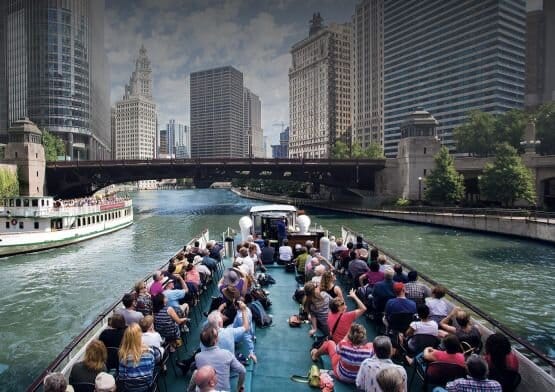 The top of a packed river cruise boat going down the river with skyscrapers surrounding river banks.