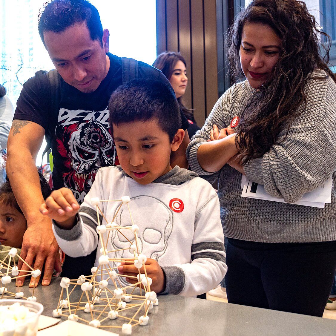 An adult couple looking on as their young son is building a project with sticks and marshmellow