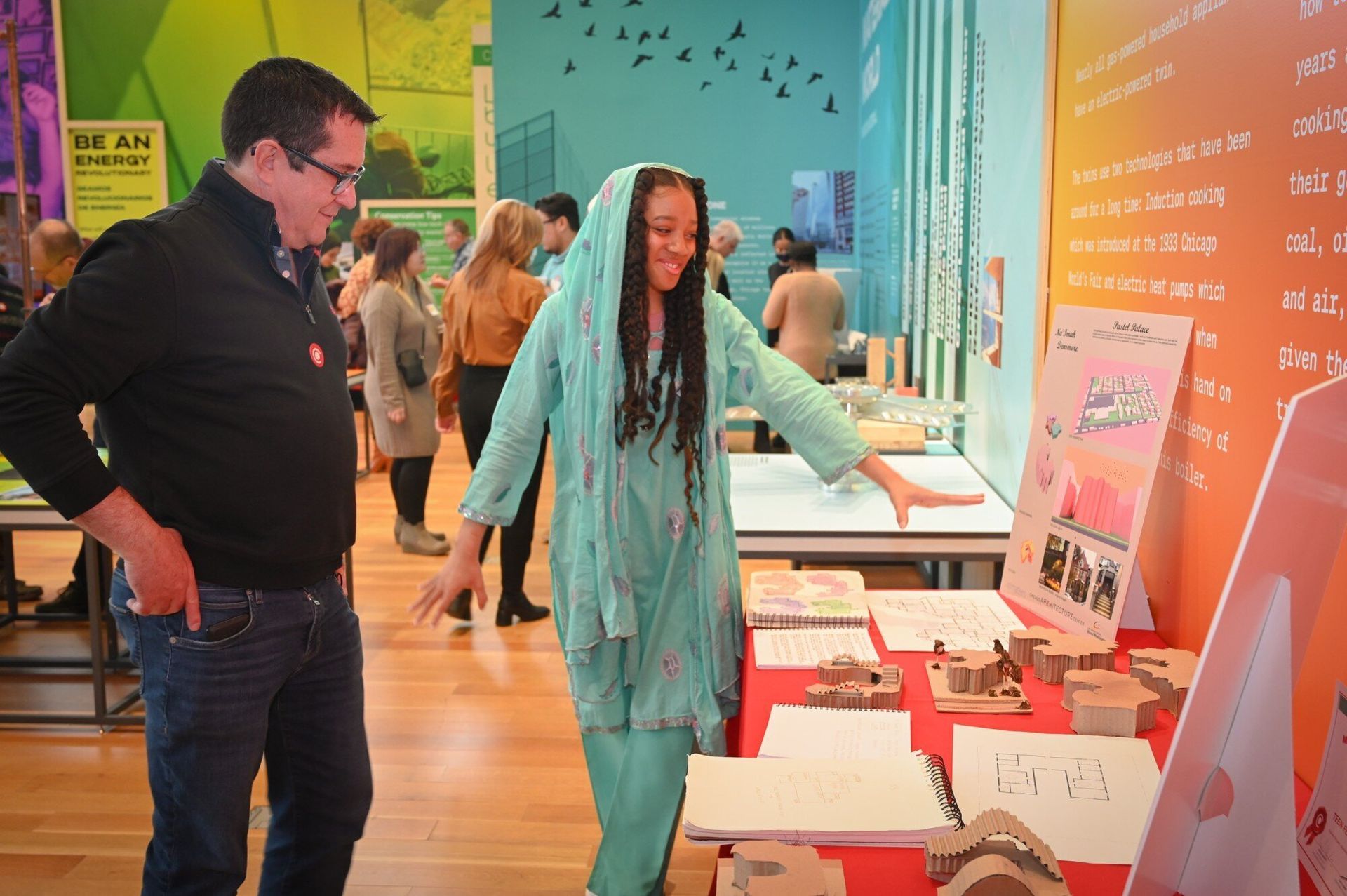A teen wearing traditional clothing showing an adult some building models on a table