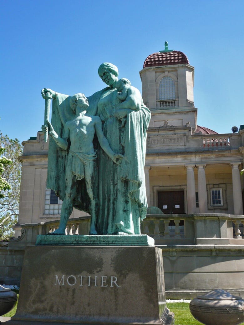 A green statue of a woman standing over a child and holding a baby, on a pedestal that reads "Mother"