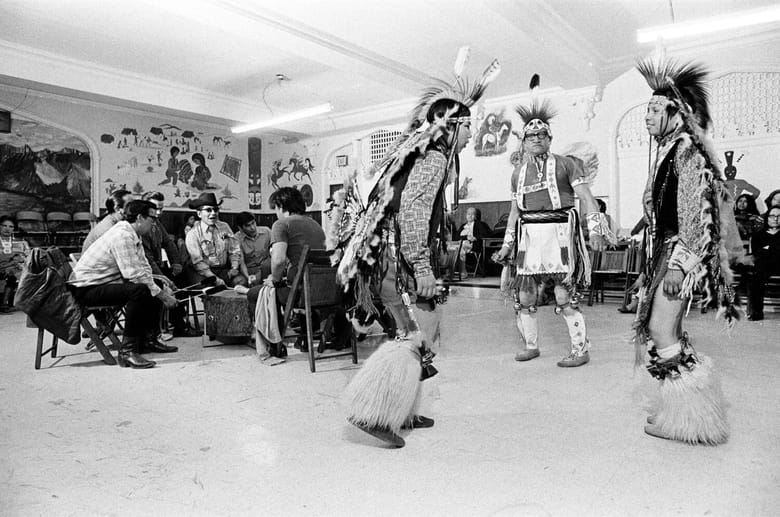 Black and white photo of 3 native american indian in a room with other people seated around a table