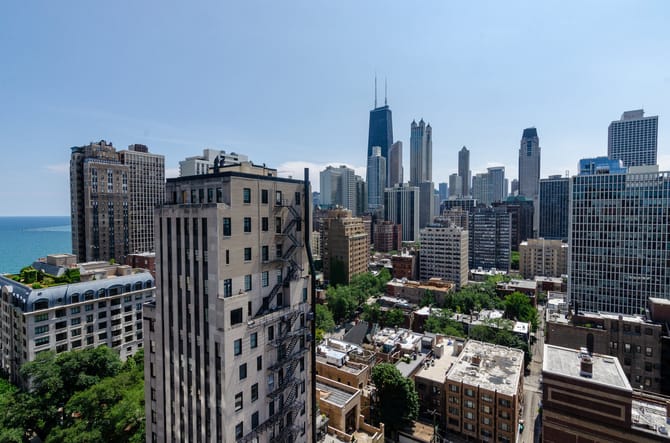 Image of the CHicago skyline looking south with a small skyscraper and some greenery in the foreground and a taller larger cluster of various buildings further away. Photo by Eric Allix Rogers.
